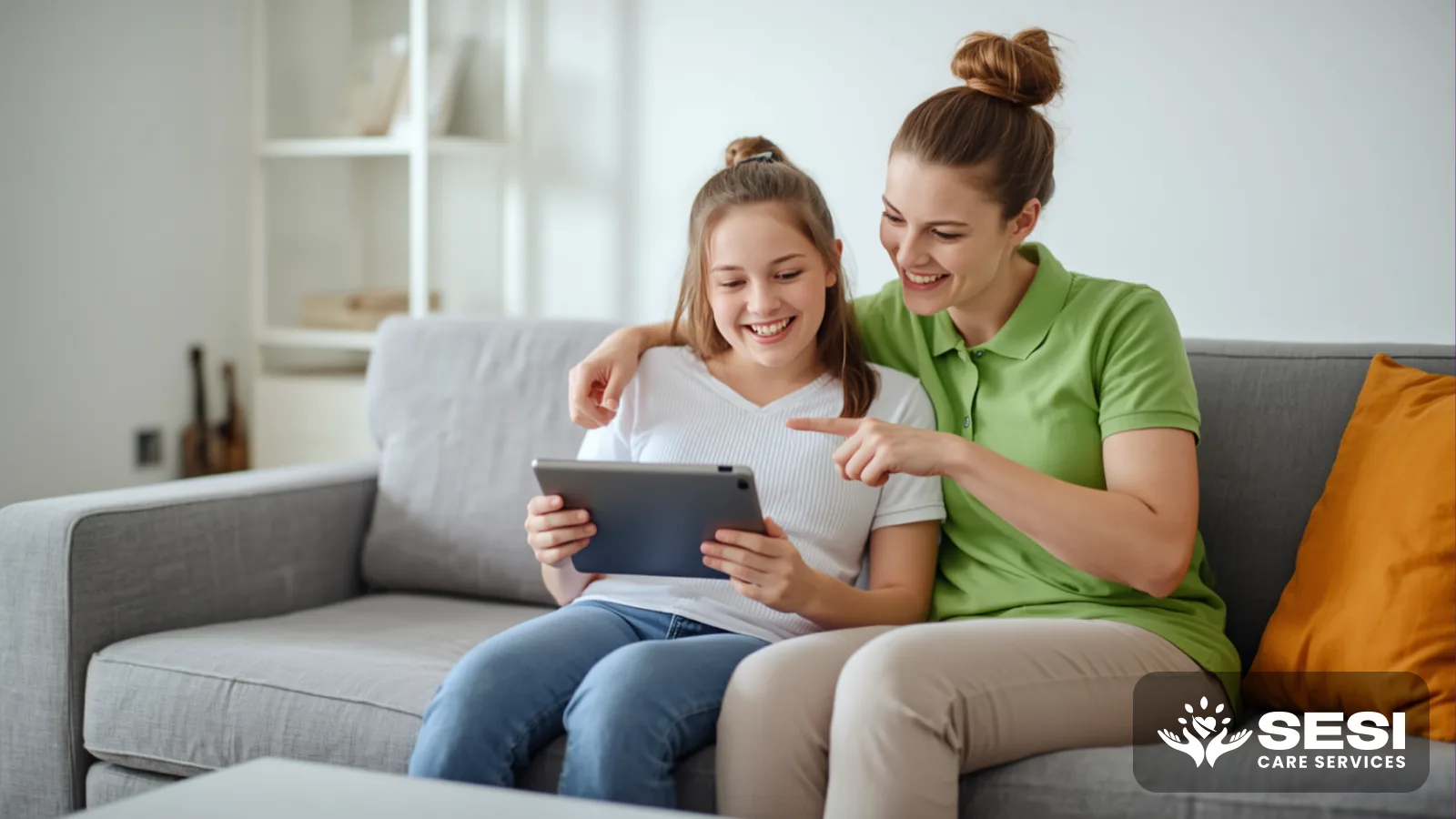 A support worker in a green polo and a young woman on a couch looking at a tablet together