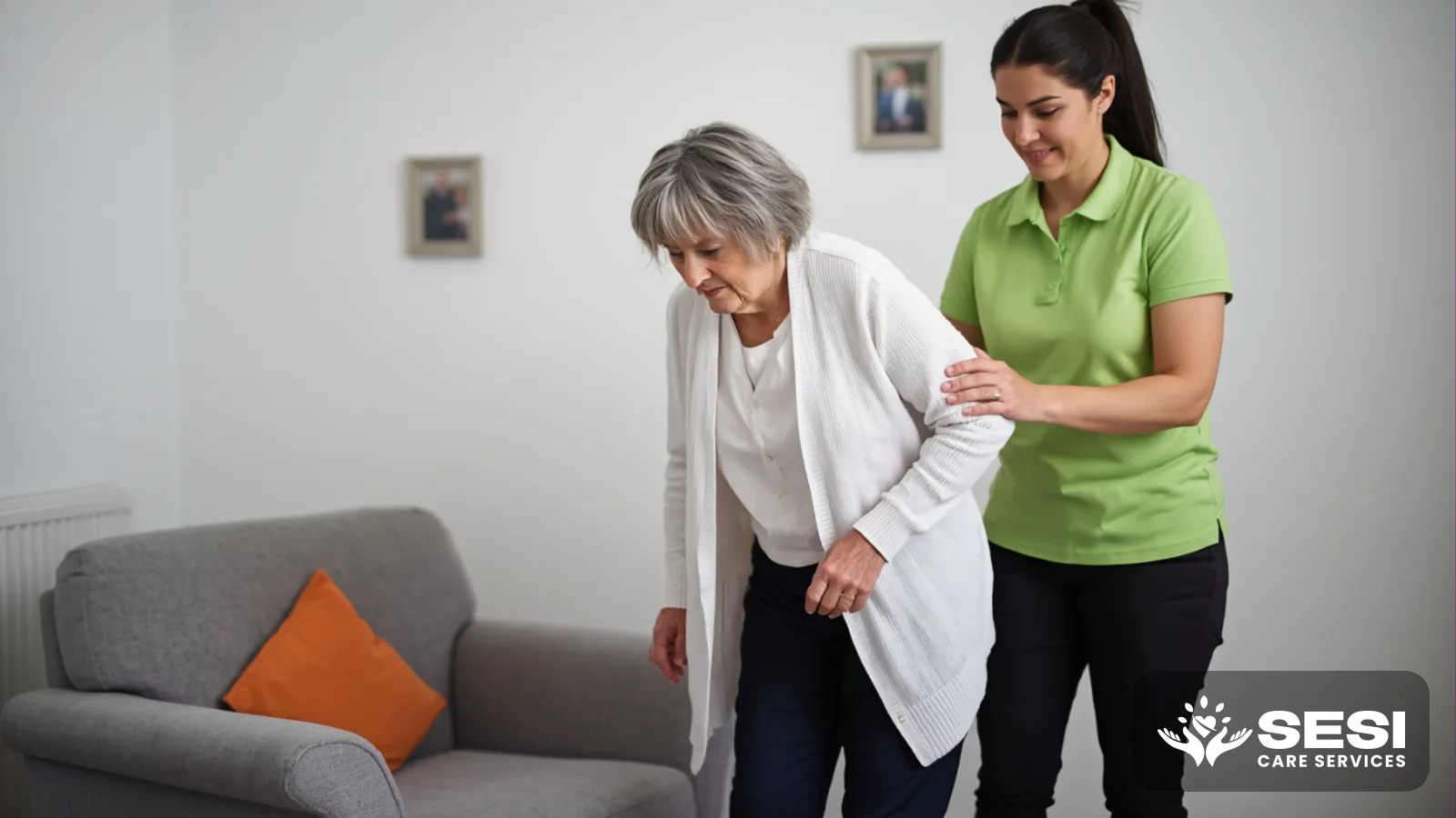 An older woman practising standing up beside a grey armchair in a white-walled lounge room while a physiotherapist in a green polo supports her elbow, an orange cushion on the chair