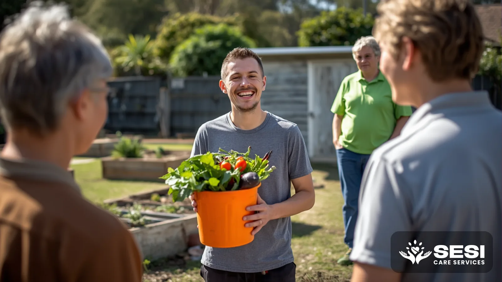 A young man holding an orange bucket of vegetables laughing with two gardeners at raised garden beds, a support worker in a green polo standing back