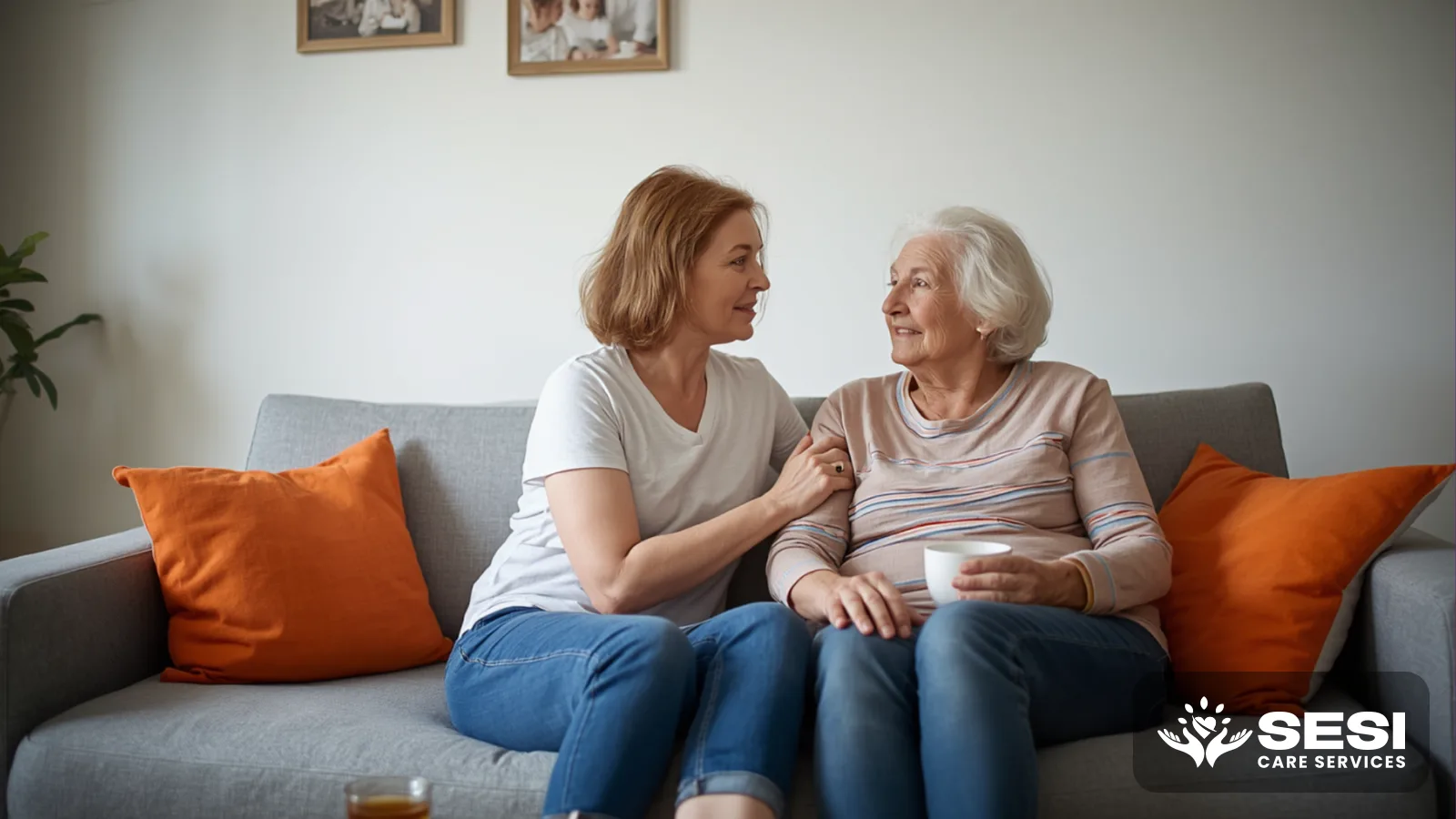 An adult daughter and her elderly mother sitting together on a grey couch mid-conversation, an orange cushion beside them and family photos on the wall