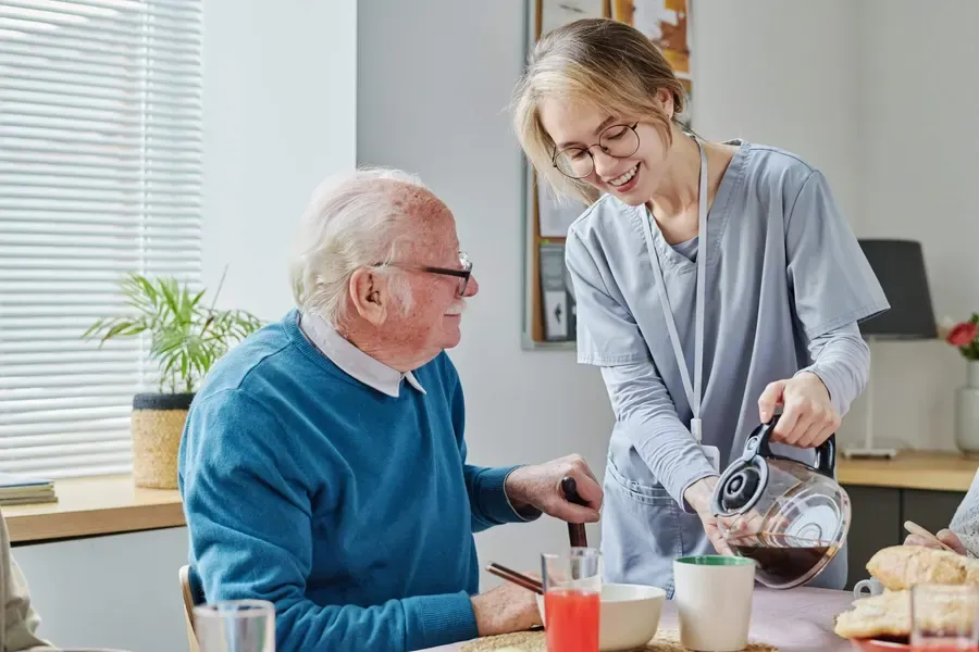 A support worker pouring a drink for an older man at his kitchen table